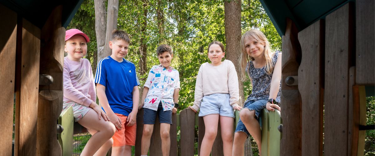 Kesävaatteisiin pukeutunut lapsijoukko seisoskelee kiipeilytelineellä ja katsoo hymyillen kameraan. Taustalla näkyy metsää.